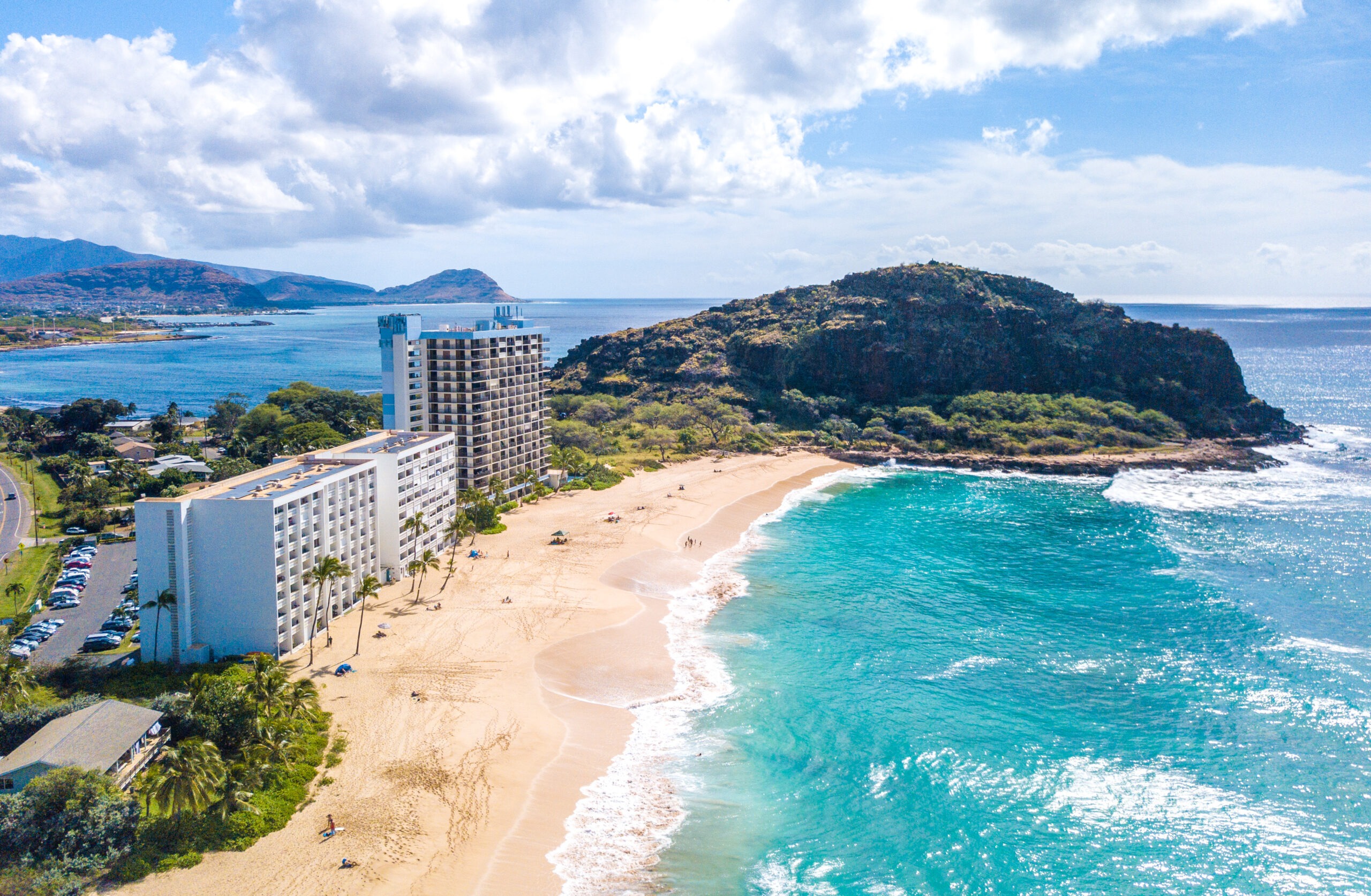 Aerial view of a tropical beach with soft sand, clear turquoise water, and hotel buildings lining the shore, under a bright blue sky.