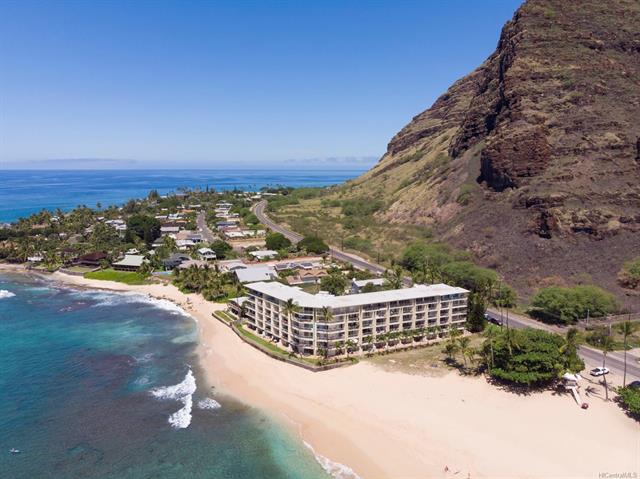 Aerial view of a beachfront resort nestled against a mountain backdrop, with clear water and a sandy beach, highlighting the scenic coastal landscape.