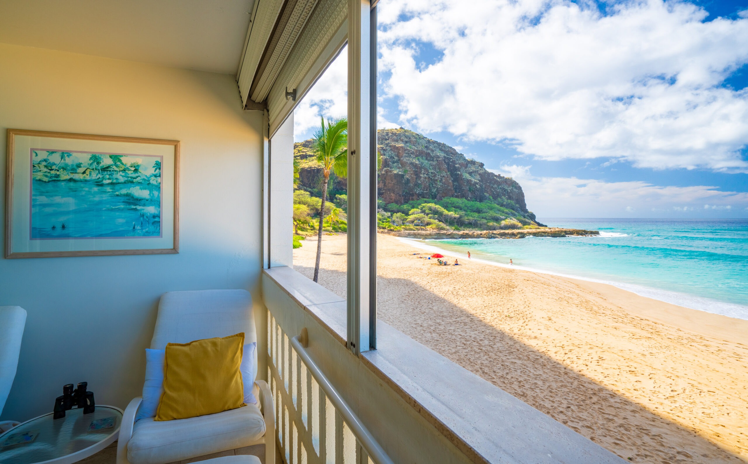 Ocean view from a balcony featuring white chairs and a yellow pillow. The beach has soft sand, gentle waves, and green cliffs in the background.