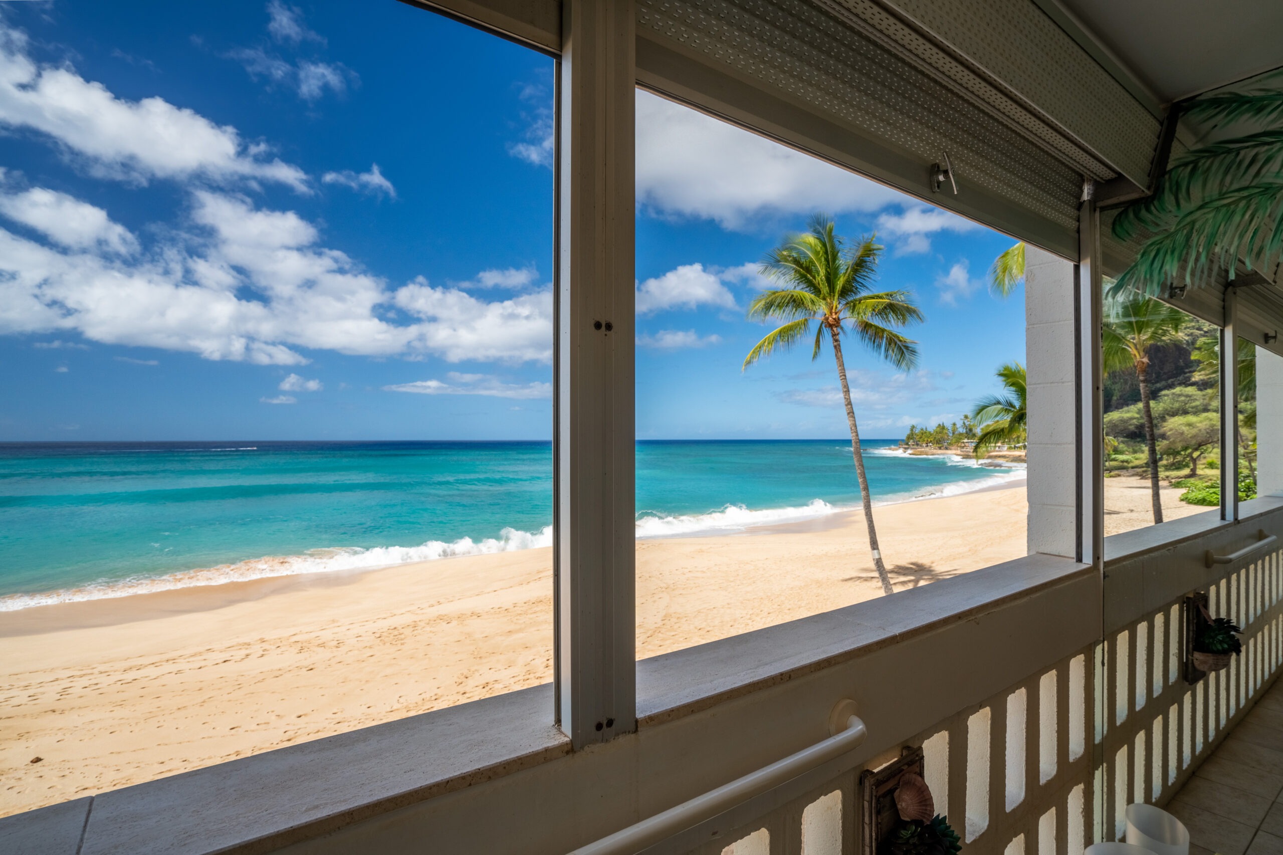 Tropical beach view from a balcony, featuring golden sand, turquoise waves, and palm trees under a blue sky with fluffy clouds.