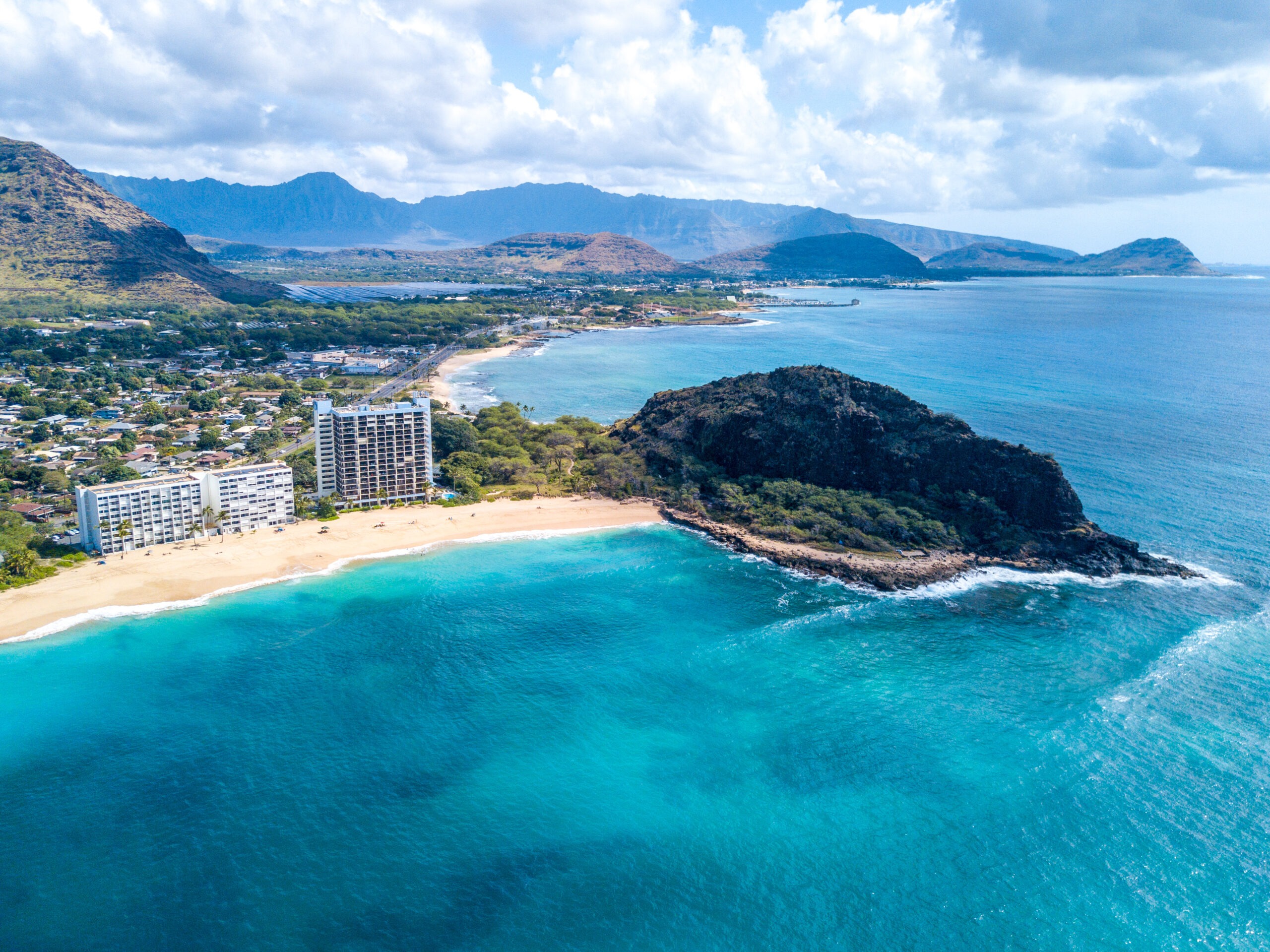 Coastal view of a sandy beach with two hotels, surrounded by turquoise waters and lush mountains, showcasing a tranquil tropical paradise.
