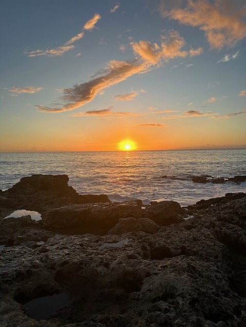 Sunset over the ocean, with vibrant orange and yellow hues illuminating the sky. Rocky shoreline in the foreground, creating a serene coastal scene.