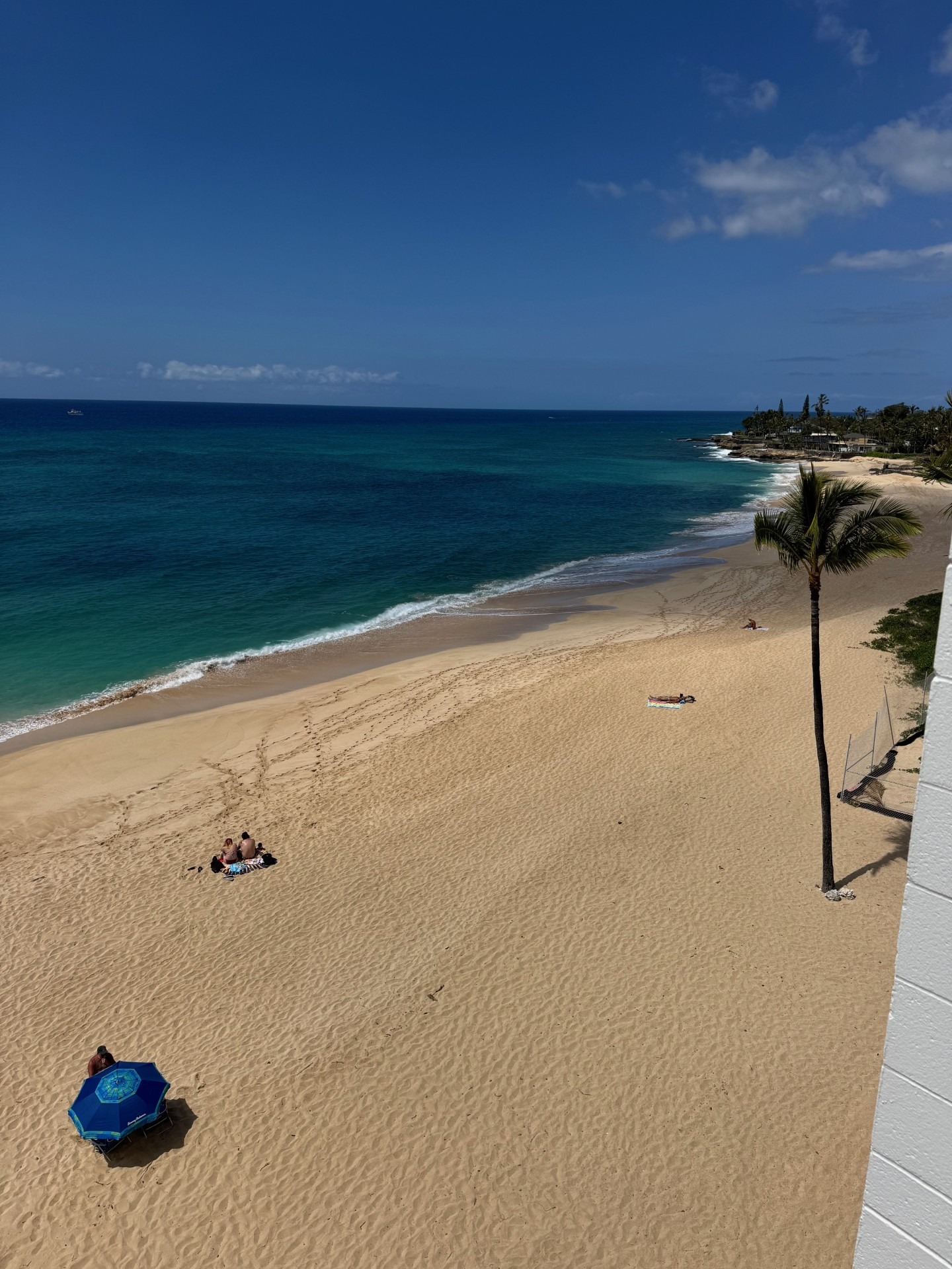 Calm beach scene with turquoise waters, golden sand, and a few sunbathers under a blue umbrella, showcasing a relaxing coastal getaway.