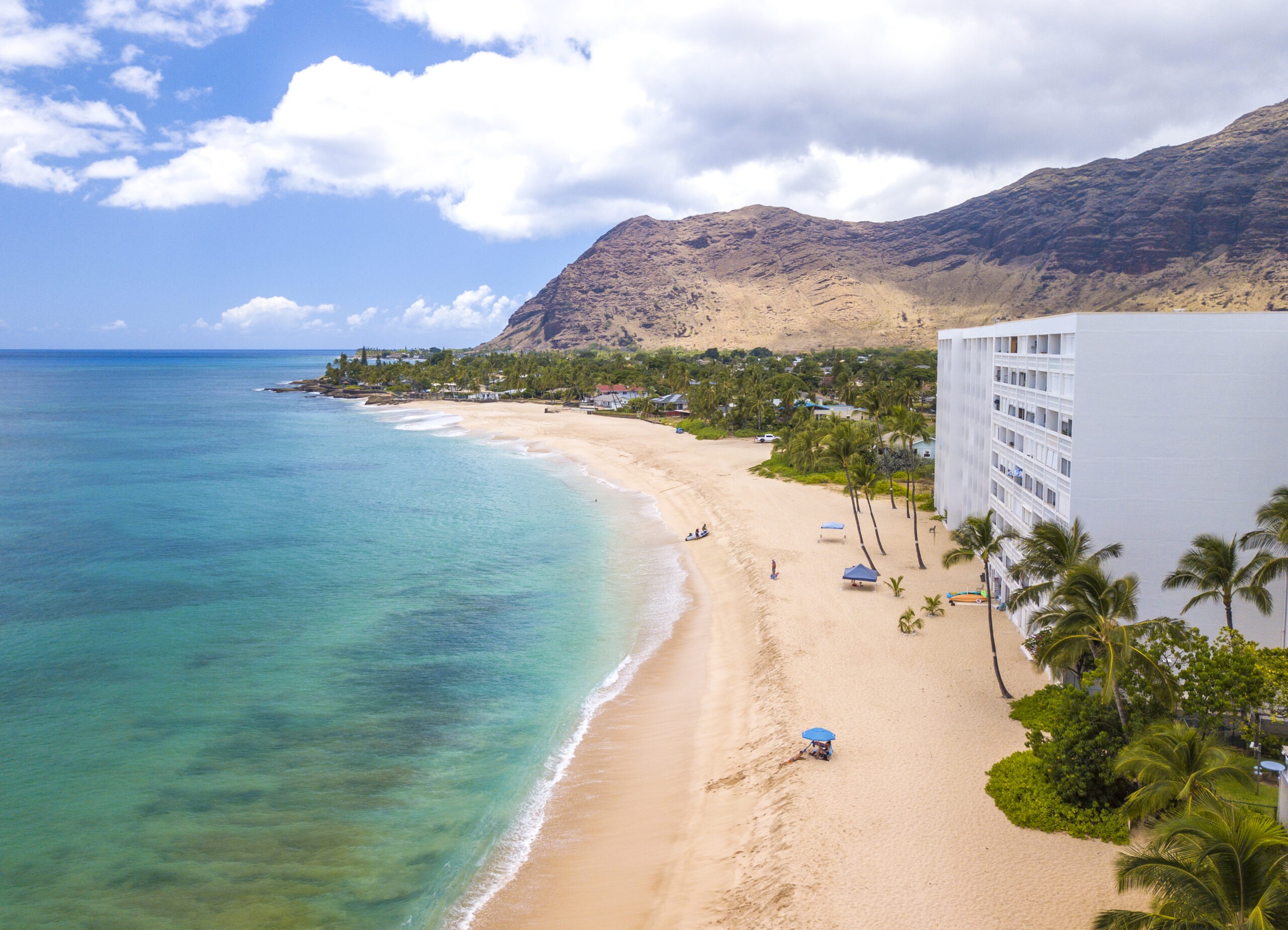 Clear blue waters gently lap a sandy beach lined with palm trees and an apartment building. Mountains rise in the background, offering a serene tropical scene.