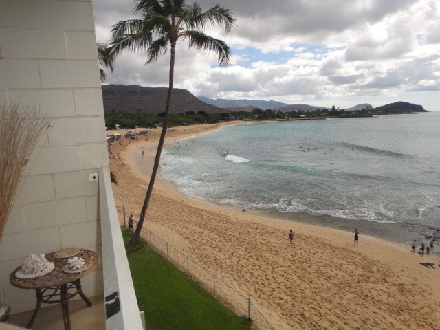 Scenic view of a beach with golden sand and gentle waves, framed by a palm tree and distant mountains, capturing a relaxing coastal atmosphere.
