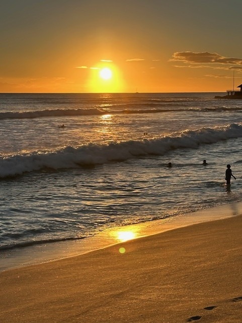 Sunset over the ocean, casting golden hues on the waves and wet sand, with silhouettes of people enjoying the shoreline.
