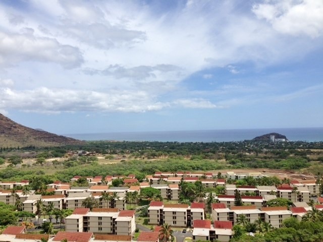 Aerial view of a coastal community near a mountain, featuring multistory buildings surrounded by lush greenery and the ocean in the background.