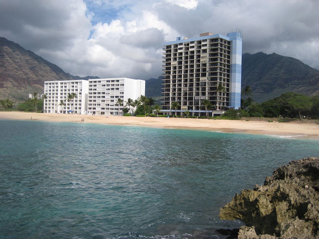 Aerial view of a coastal area featuring low-rise buildings with red roofs, lush greenery, and a distant mountain range under a partly cloudy sky.
