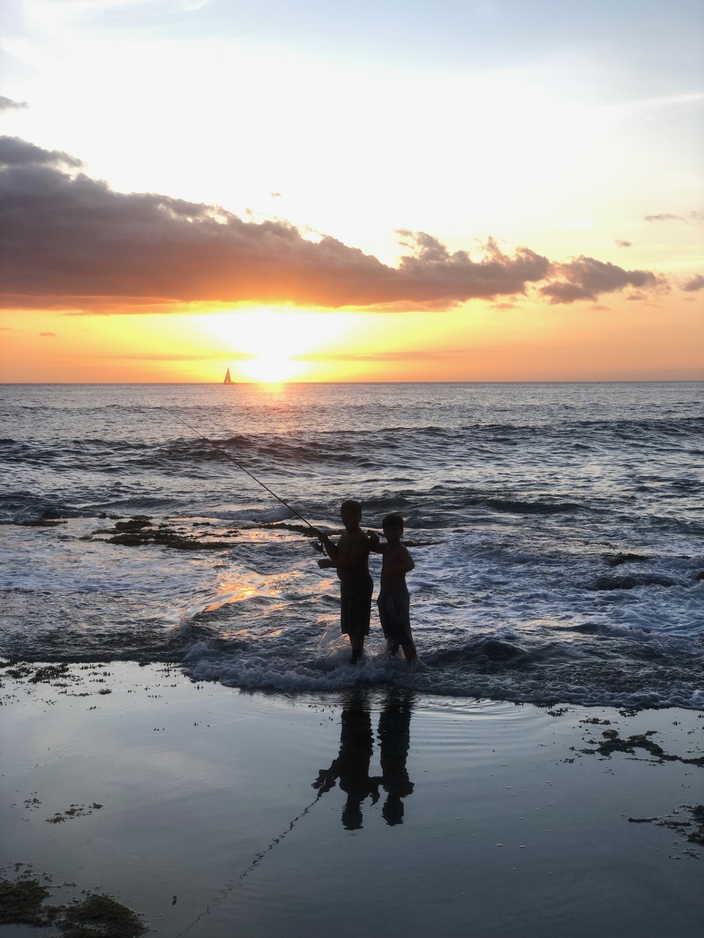 Two boys standing in the shallow water of the beach, fishing at sunset. Their silhouettes are reflected on the water as the sun sets behind them, with a sailboat visible in the distance.