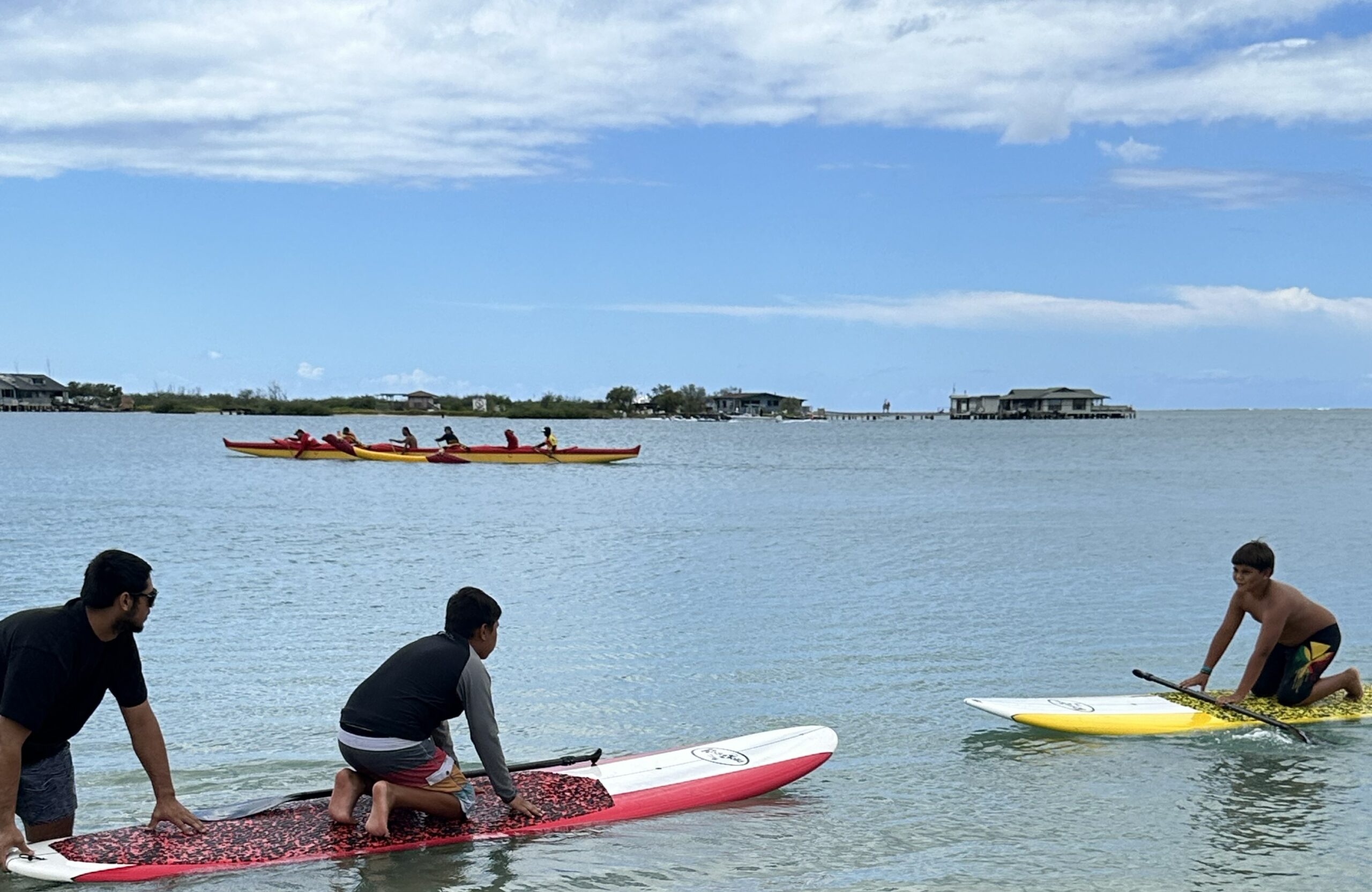 Two boys kneeling on stand-up paddleboards in the shallow water, one with a paddle. In the background, a group of people is kayaking on the water with houses and a pier in the distance.